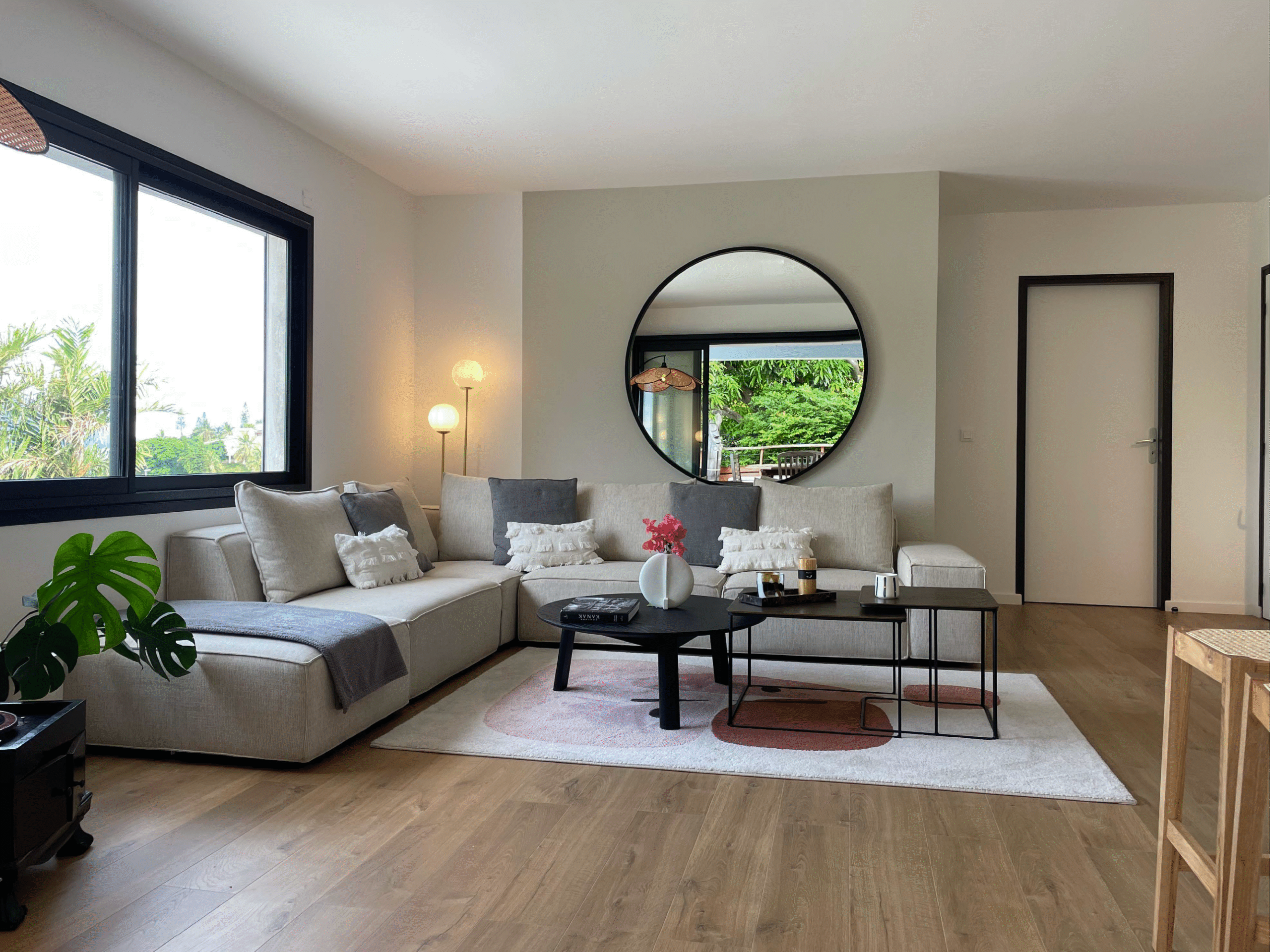 Salon contemporain avec grand canapé beige, miroir rond et baie vitrée offrant une vue sur la mer, réalisé par Grichka Interiors Design