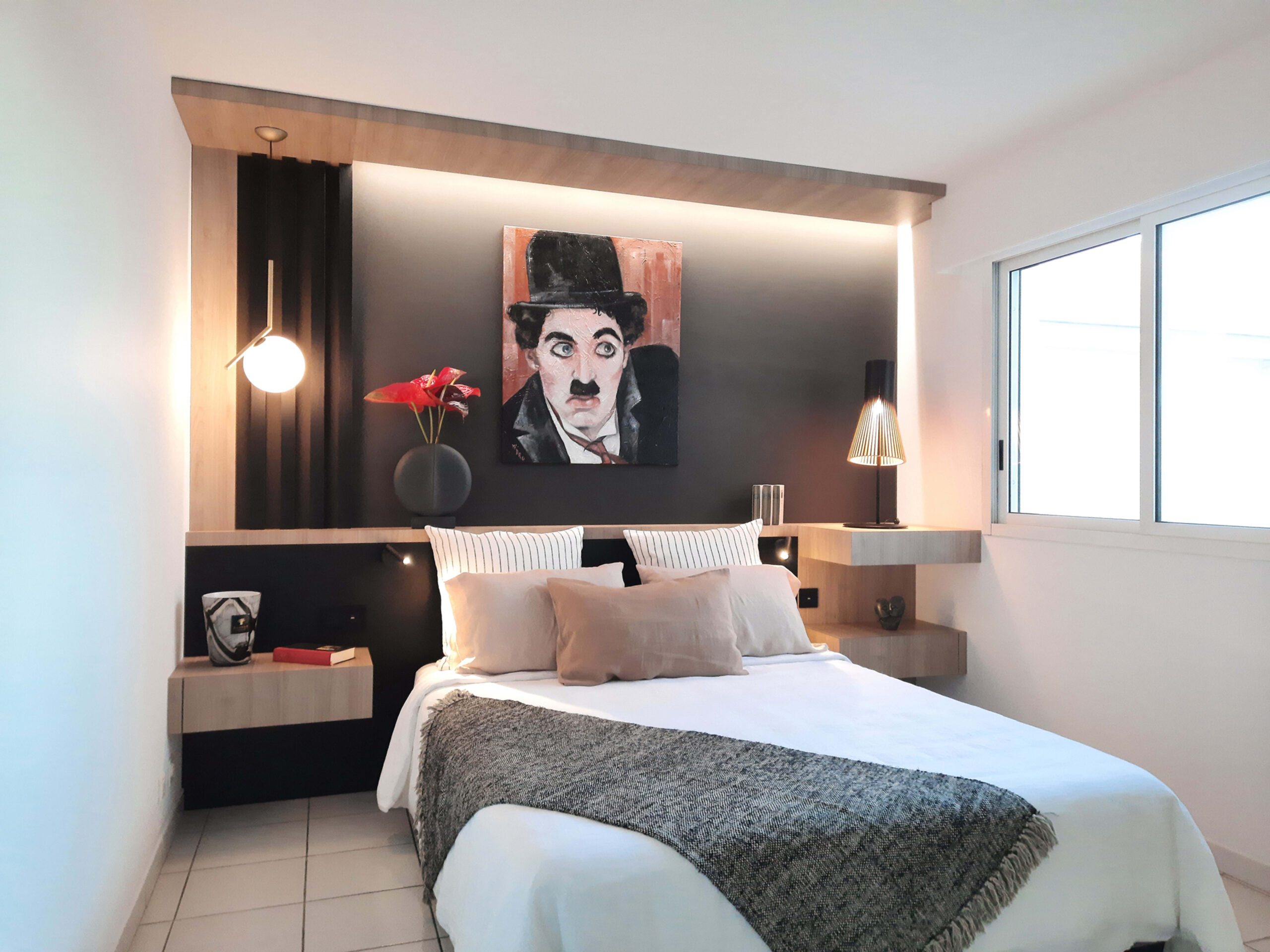 Chambre moderne avec mur noir, tête de lit en bois clair, éclairages design et tableau de Charlie Chaplin, réalisée par Grichka Interiors Design