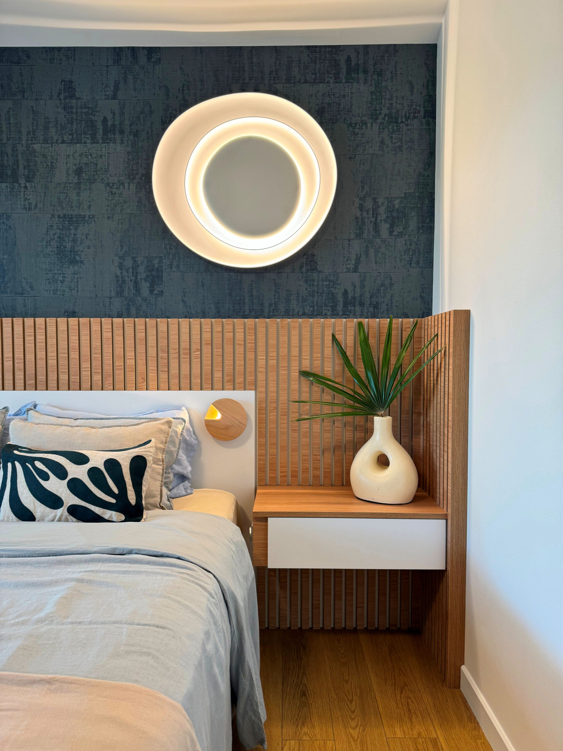 Chambre design avec tête de lit en bois rainuré, mur bleu texturé et luminaire mural circulaire, réalisée par Grichka Interiors Design