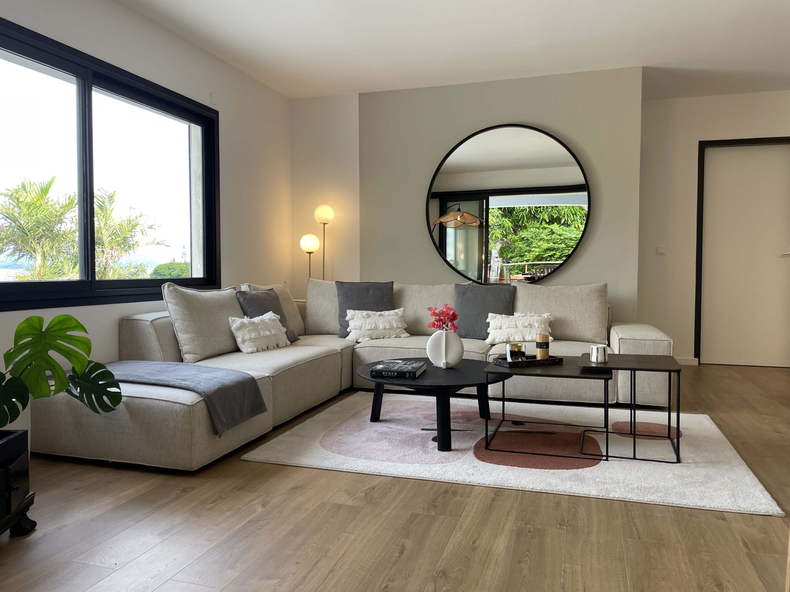 Salon contemporain lumineux avec canapé d’angle beige, tables basses noires, miroir rond mural et décoration épurée, aménagement intérieur par Grichka Interiors Design.